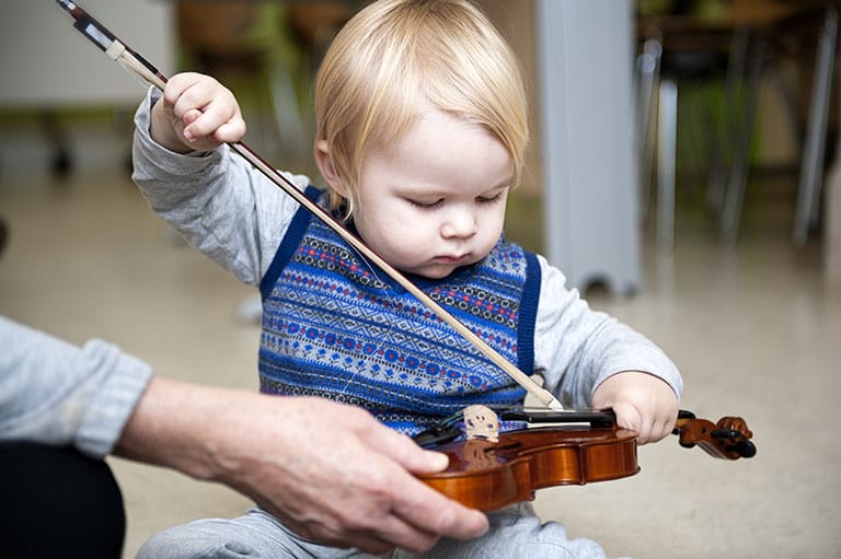 A toddler holds a violin on his lap and practices using a small bow. He is wearing a knitted blue sweater vest.