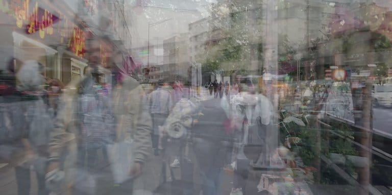 A busy, double-exposed photograph of a bustling city. People walk in different directions, overlayed with motion shots of traffic, shops and blocks of flats.