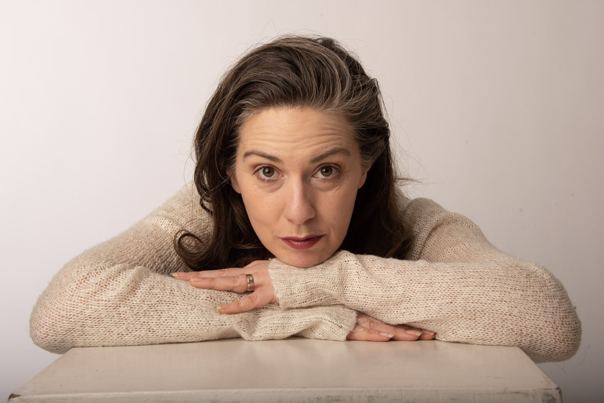Comedian Meryl O'Rourke poses leaning forward with her chin atop her folded hands. She is wearing a cream sweater against a cream background. She has mid-length brown hair and brown eyes.