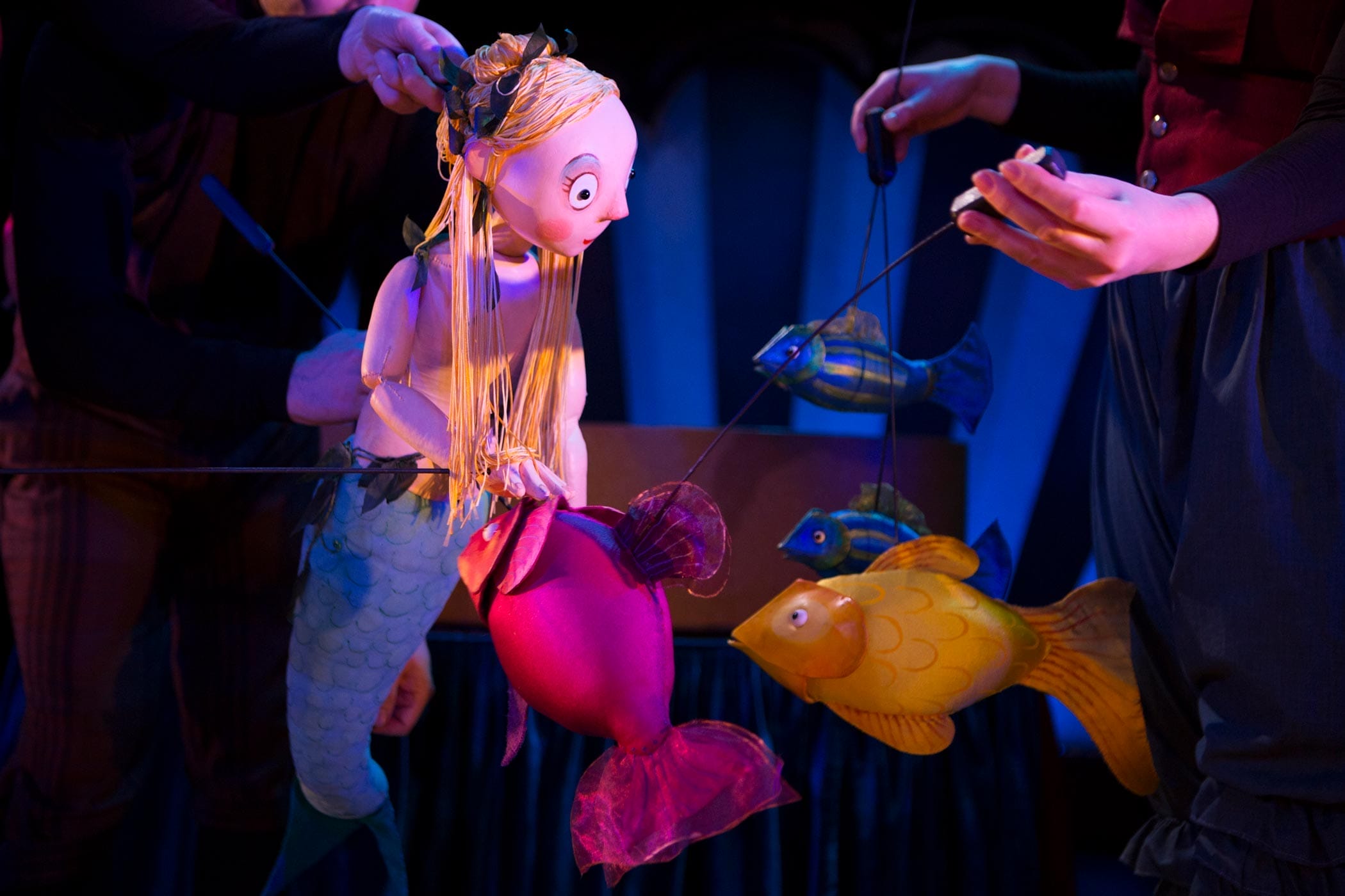 A mermaid puppet surrounded by wooden fish puppets. The hands of puppeteers can be seen.