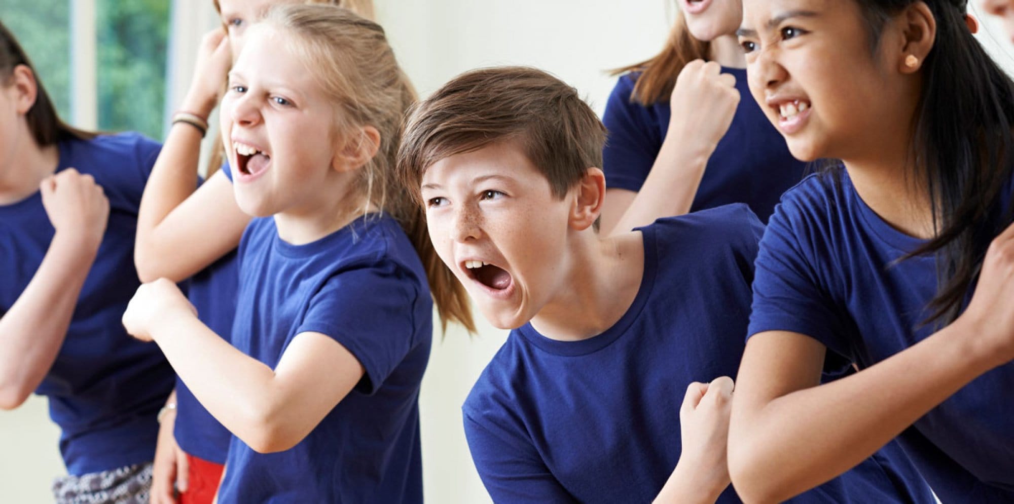 A group of young people in matching blue shirts perform musical theatre. Their mouths are open in song as they all co-ordinate their arm movements as part of their choreography.