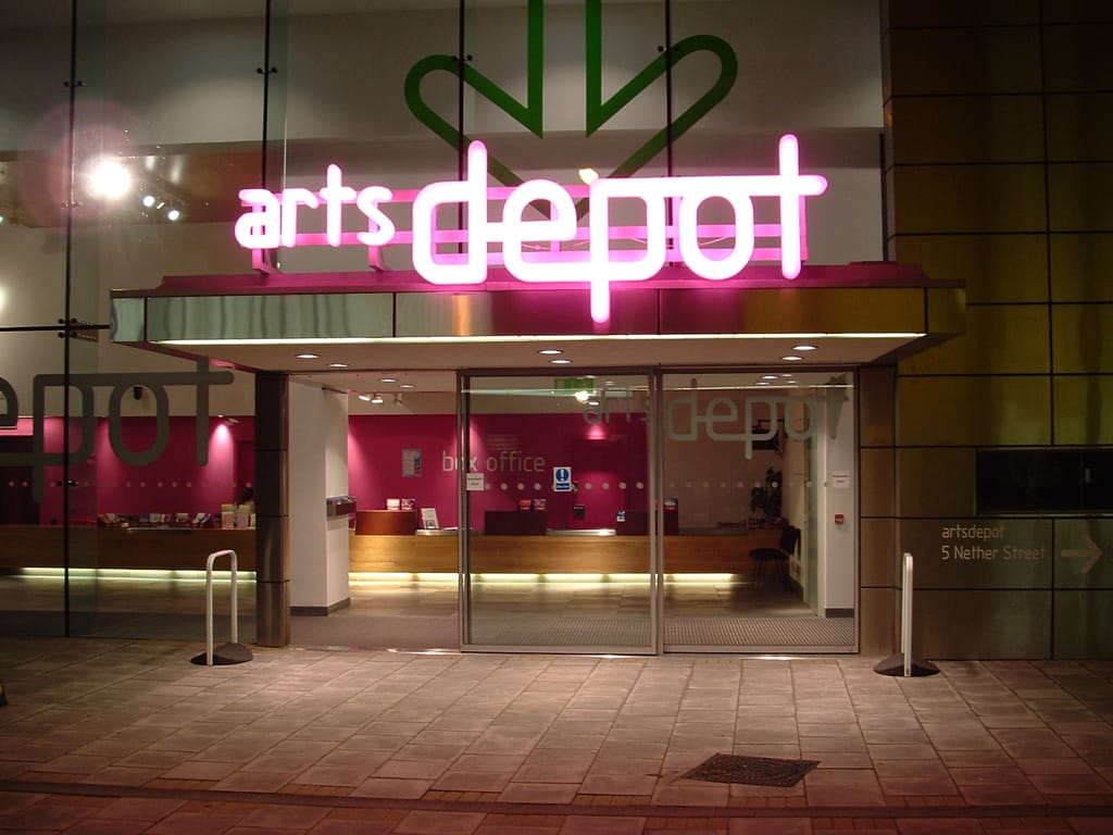 The exterior of artsdepot, pictured at night. A bright pink neon sign reads artsdepot over the entrance. The Box Office counter can be seen inside.