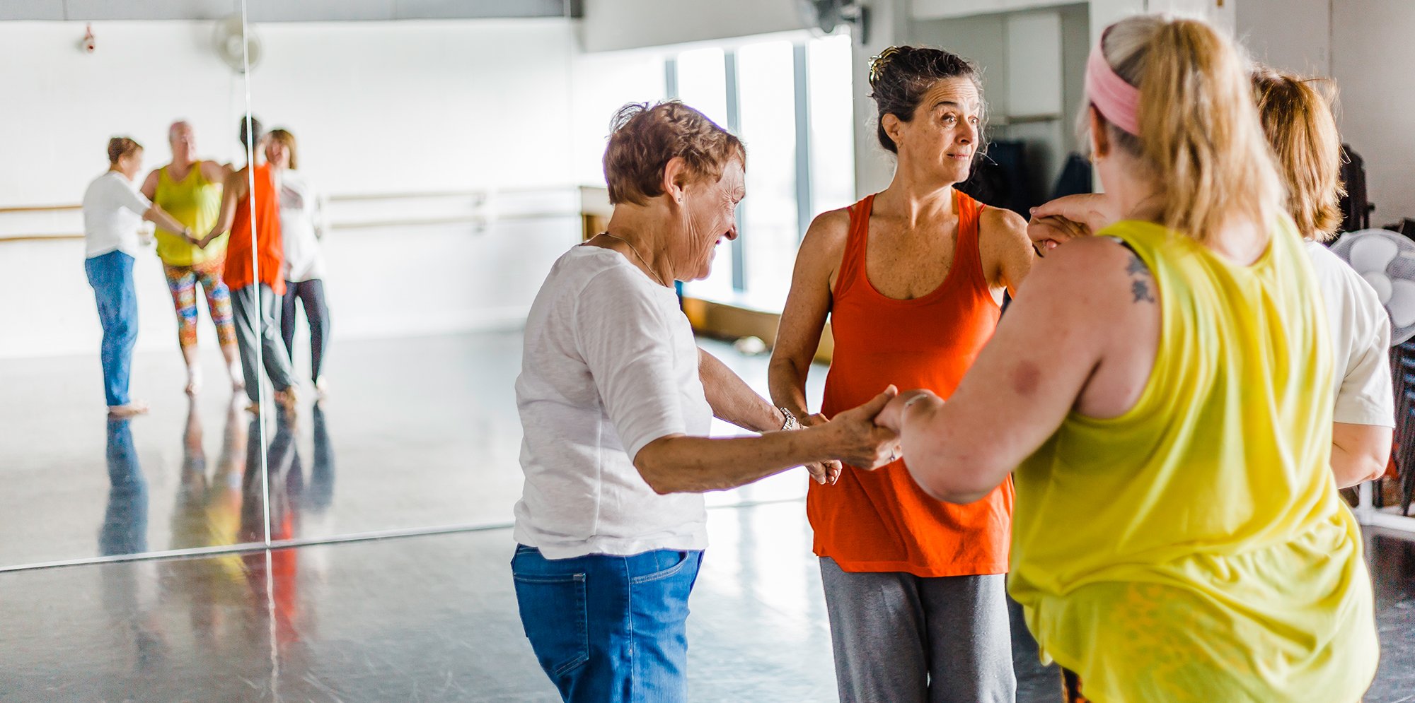 Members of the over 55s dance group warm up in the studio. They are wearing vibrant dance wear: bright yellow and orange tops. We see the group reflected in the large dance studio mirror.