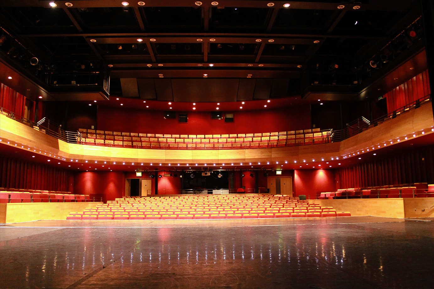 A high quality photograph of artsdepot's Pentland Theatre auditorium, photographed from the stage, offering an expansive view of the seating. The auditorium is dark red with wooden seating. Stage lights illuminate the image.