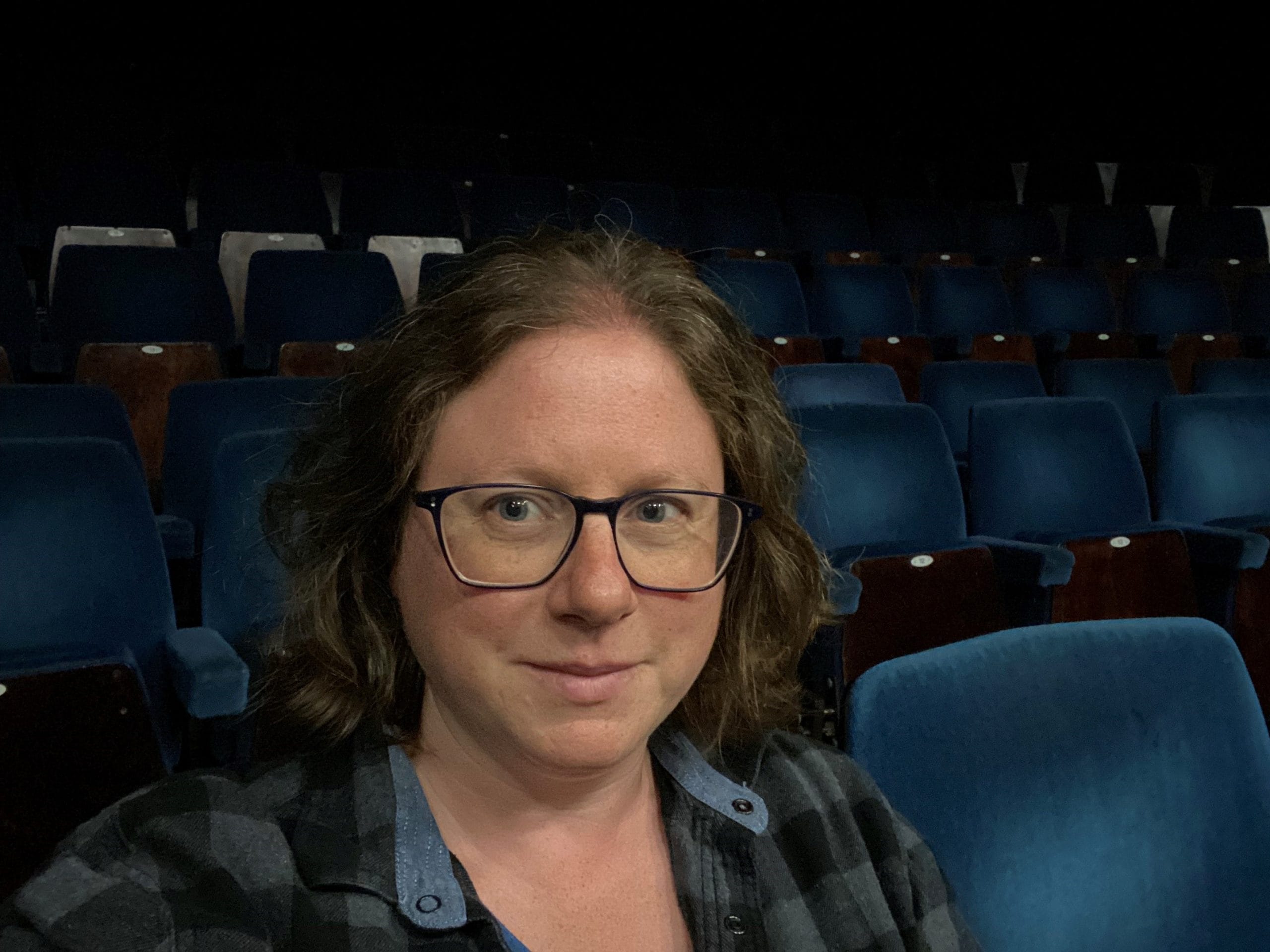 Rachel Barnett Jones poses for a selfie in a theatre auditorium. The seats behind her are blue velvet. Rachel faces the camera with a smile. She has short-mid length brown hair and is wearing glasses and a blue plaid shirt.