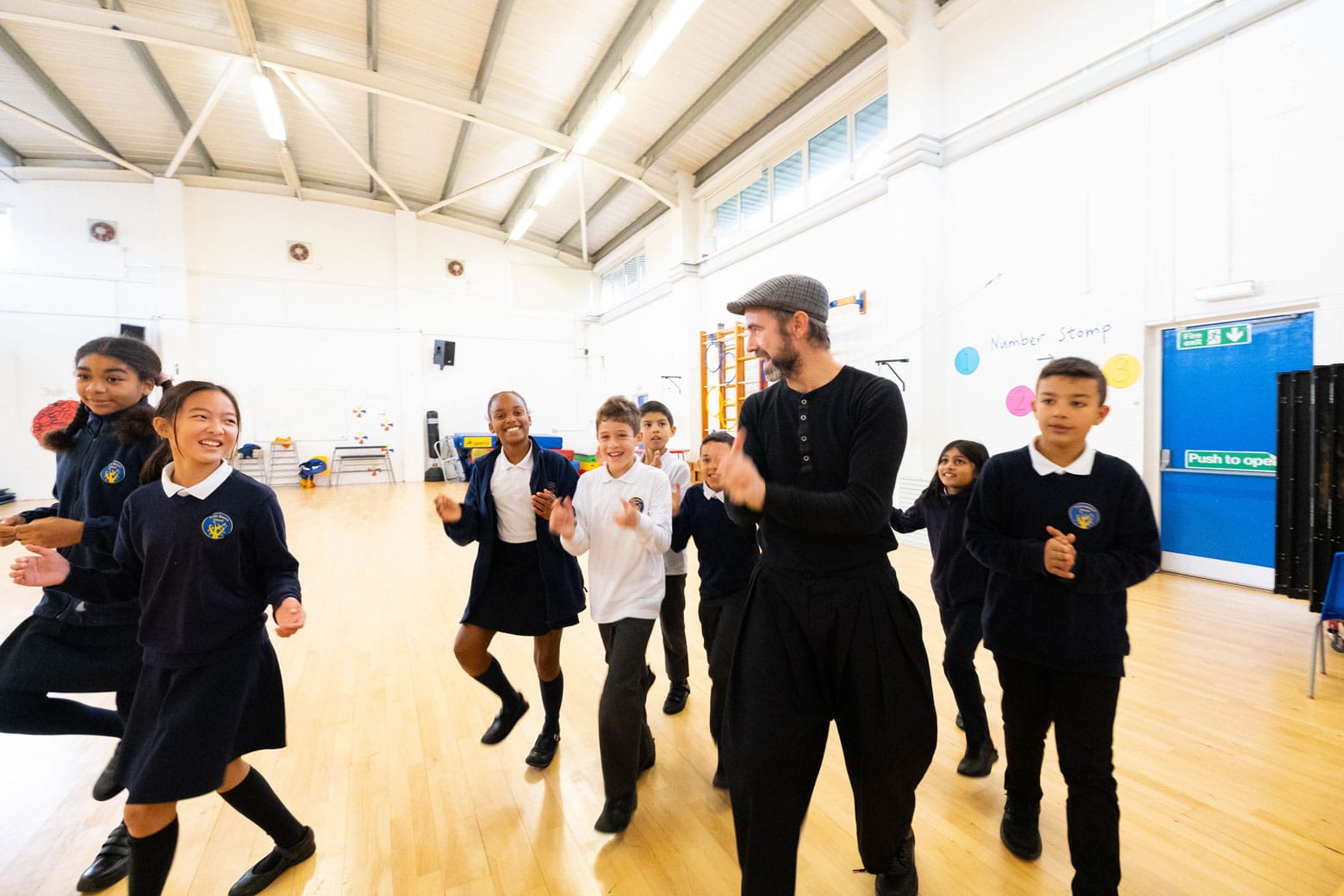 A group of school pupils doing a workshop in a school hall. Eight smiling pupils in uniform follow the workshop leader, a bearded man wearing a cap. The group are smiling and clapping.