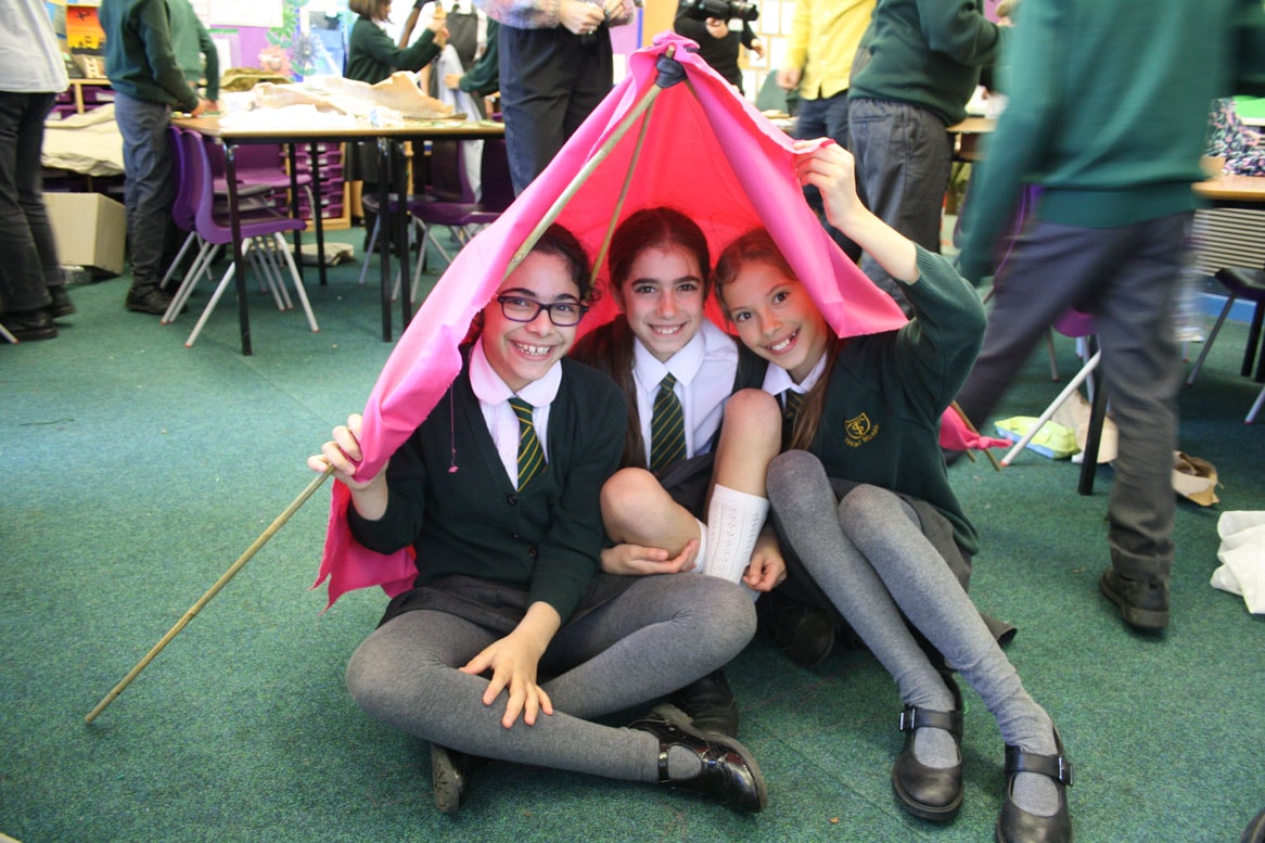 Three smiling students sit cross legged under a structure they have made from pink fabric and sticks.
