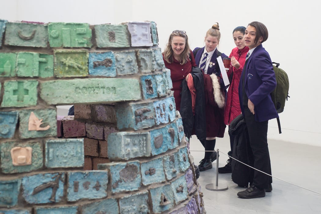 Two teachers and two students next to The Storytelling Igloo, a structure made from coloured concrete blocks, on display in a white-walled gallery.