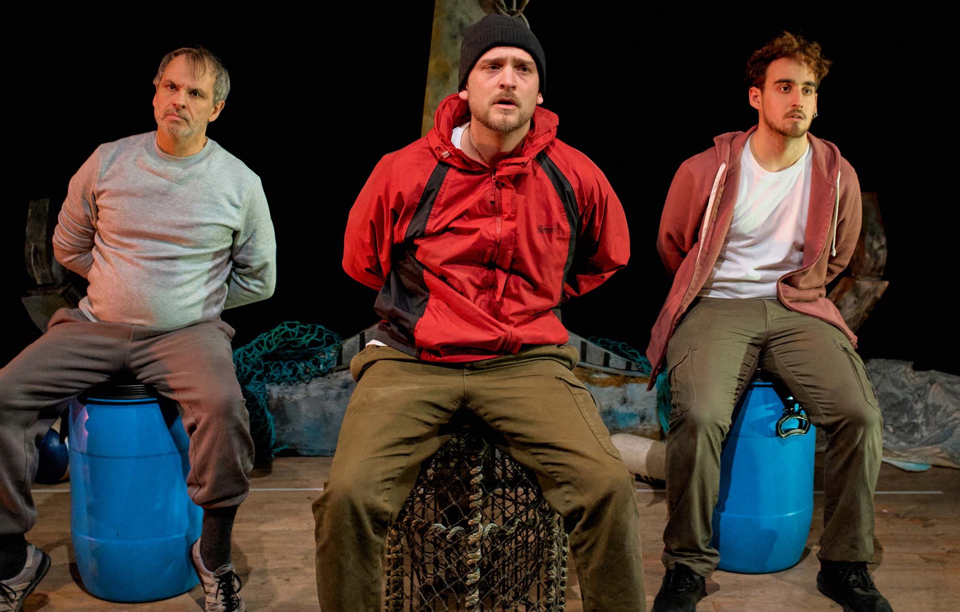 Three men sit on fishing pots and buoys with their hands behind their backs.
