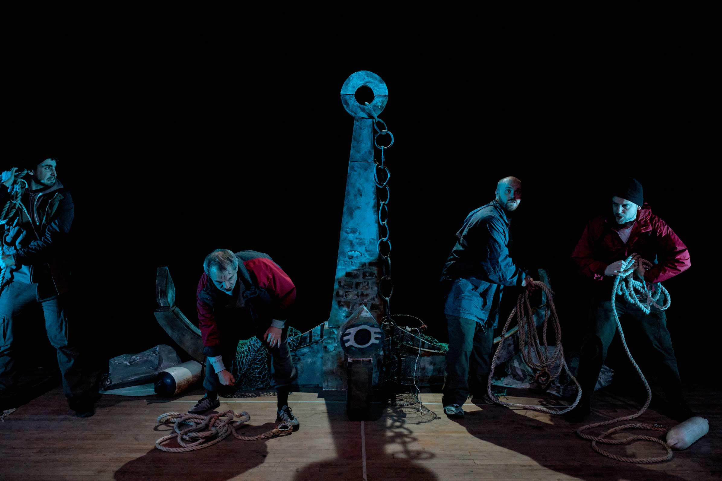 Performers on stage in The Freshwater Five - four performers gather ropes on a dimly lit stage. A large anchor can be seen behind them.