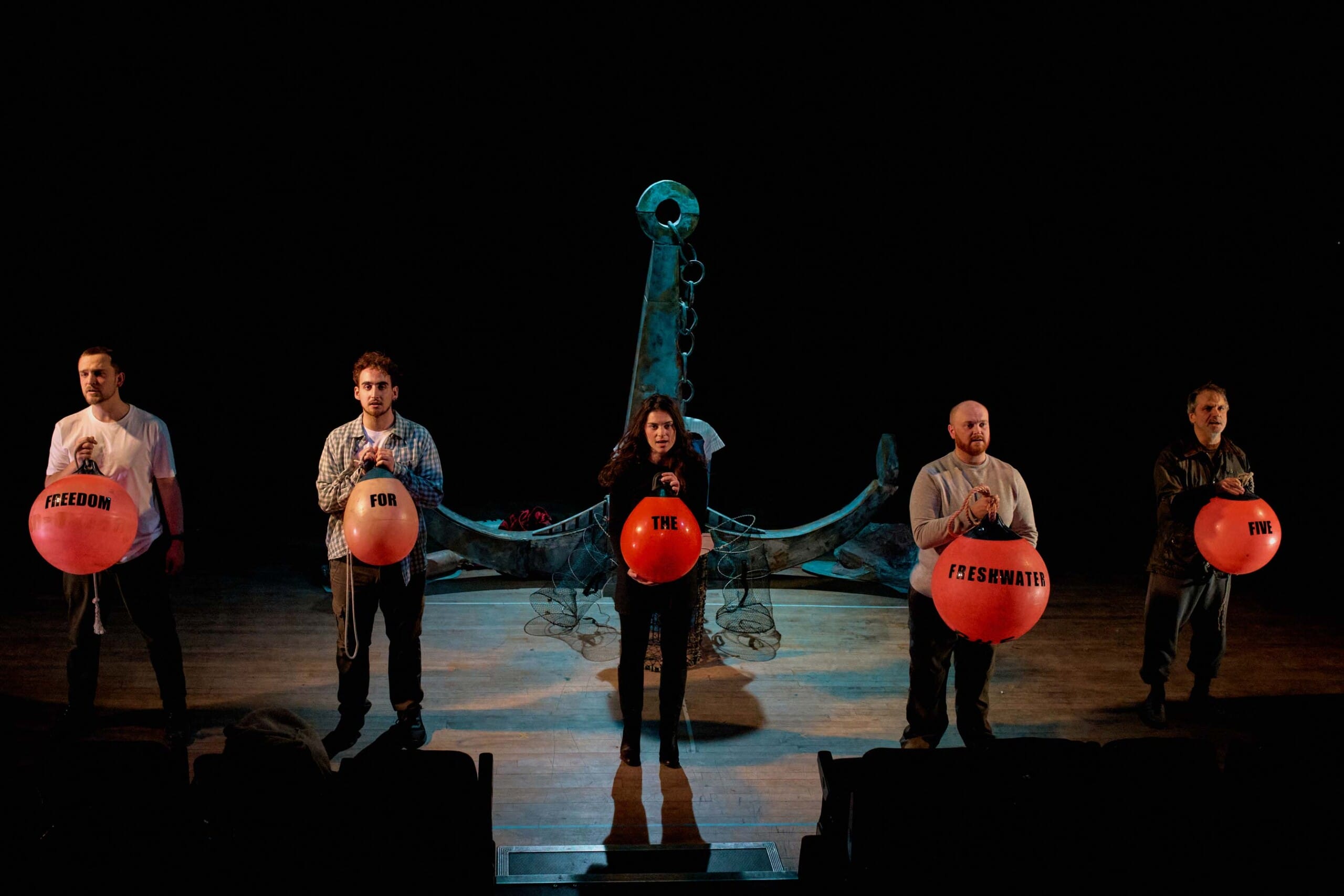 Performers on stage in The Freshwater Five - they stand in a line holding orange buoys on which black text reads Freedom for The Freshwater Five.