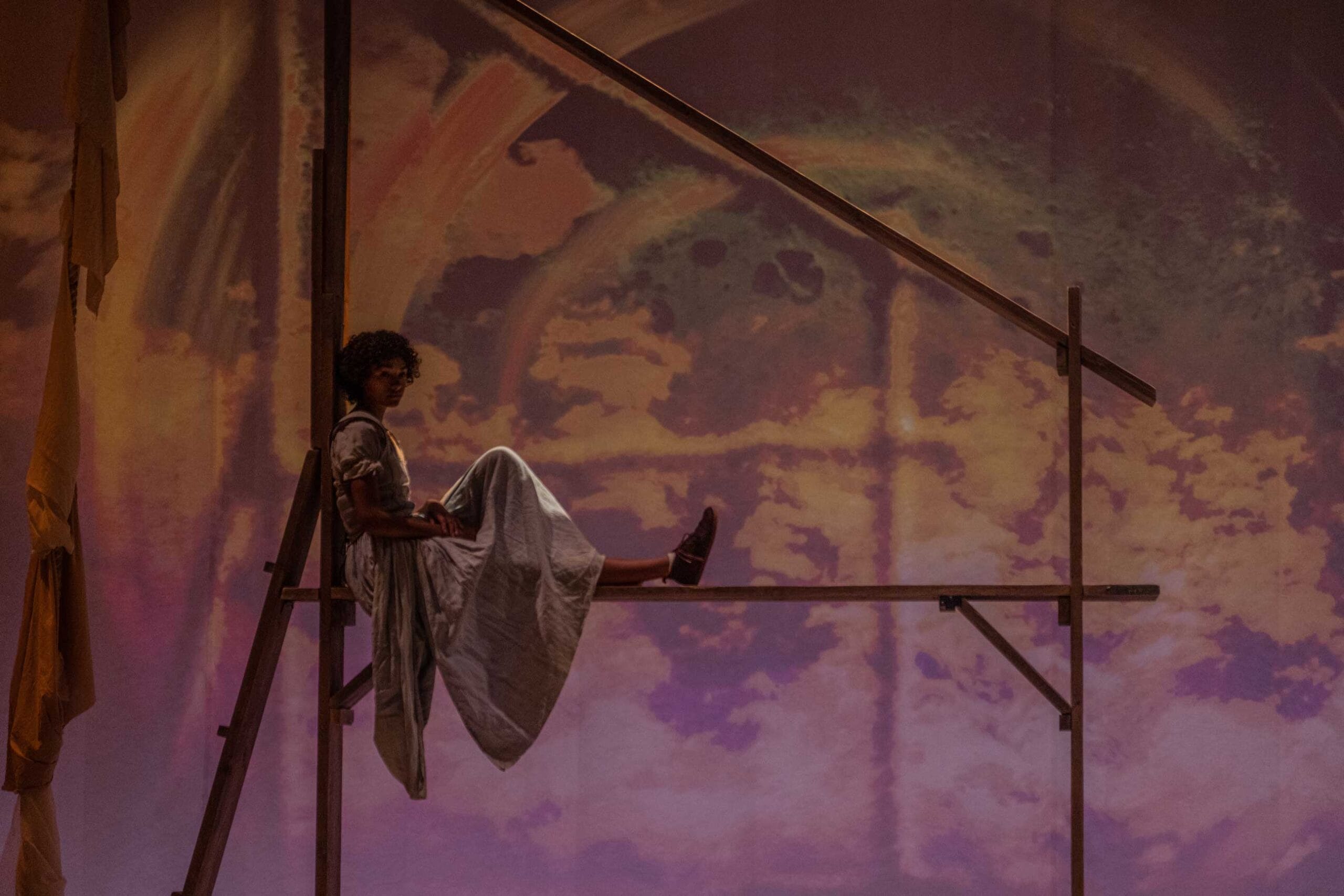 A performer sits high above the group in a thin wooden structure. She is looking into the distance and her flowing skirt hangs below her. Behind her is an abstract projection in muted colours.