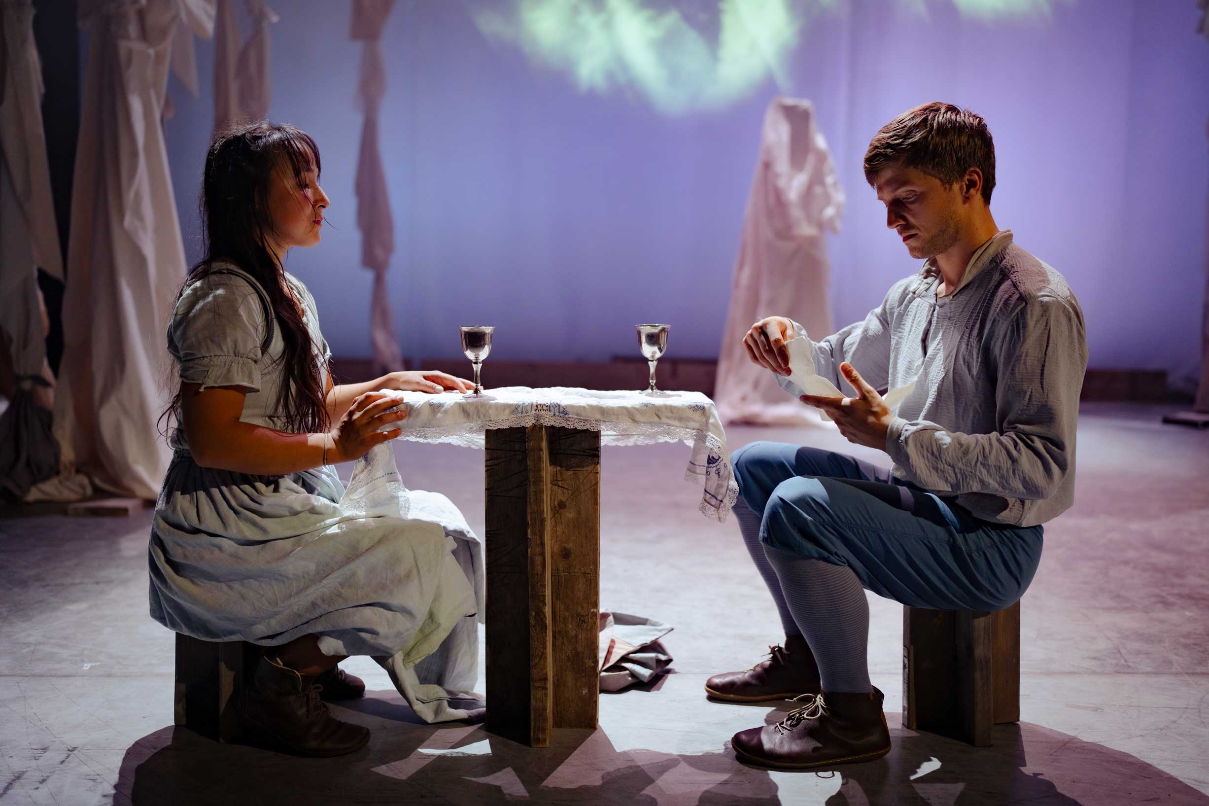 A male performer and a female performer sit at a small table covered with a white cloth and two small shiny glasses. The male performer reads from a letter.