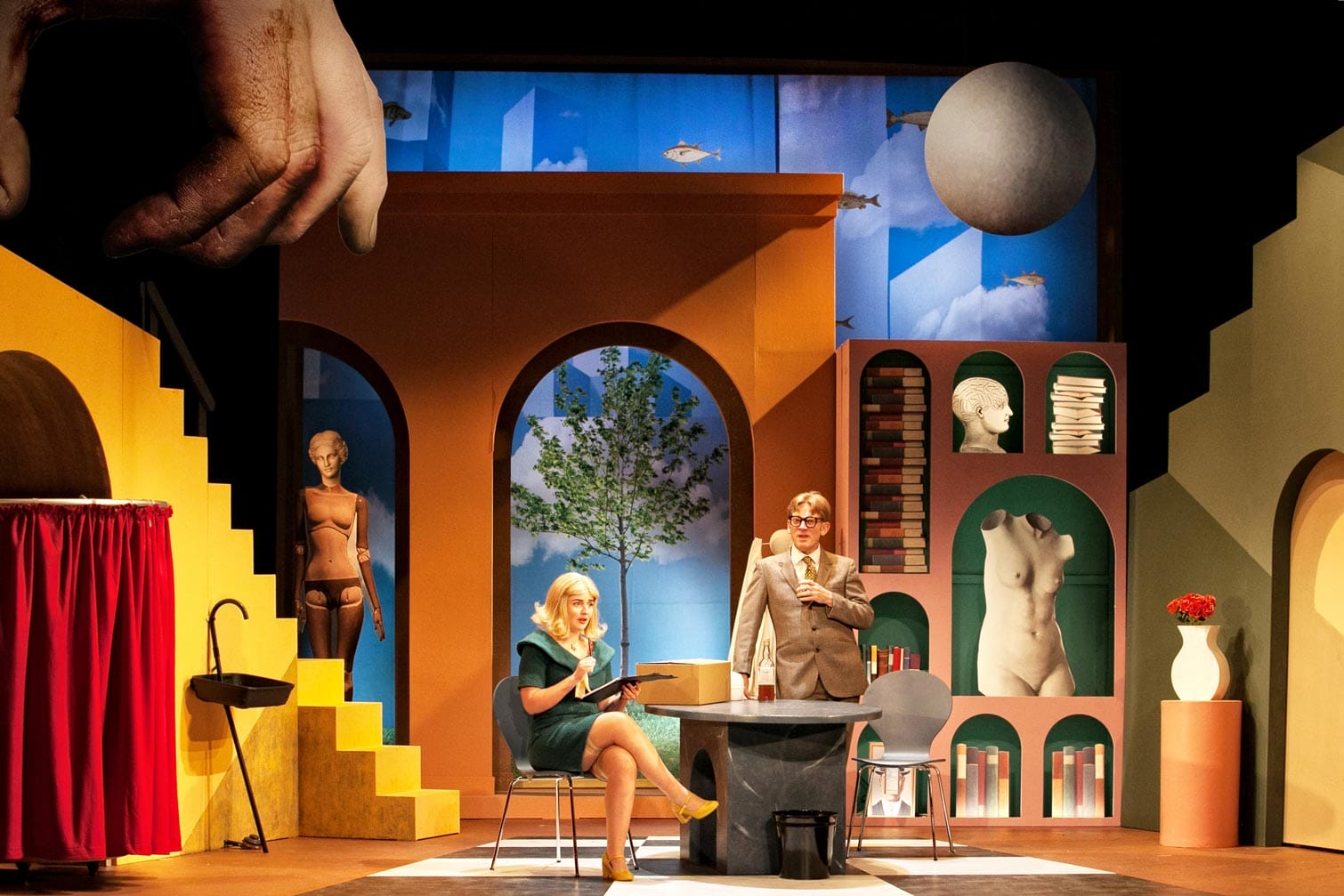 Two performers on stage in What the Butler Saw. The set is 1960s in style with elements of surrealism - there are staircases that don't go anywhere, a large realistic hand reaching down from above, and in the background a painted sky backdrop with geometric shapes and fish on it.