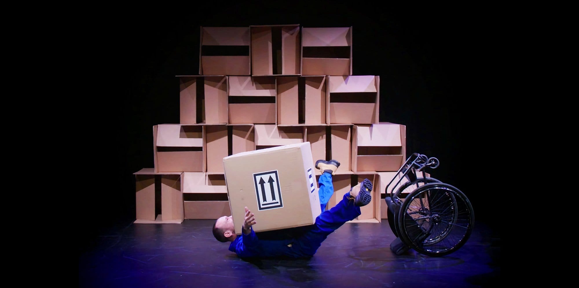 A man wearing a blue jumpsuit laying on the floor with a cardboard box ontop of him. Behind is a wall of cardboard boxes