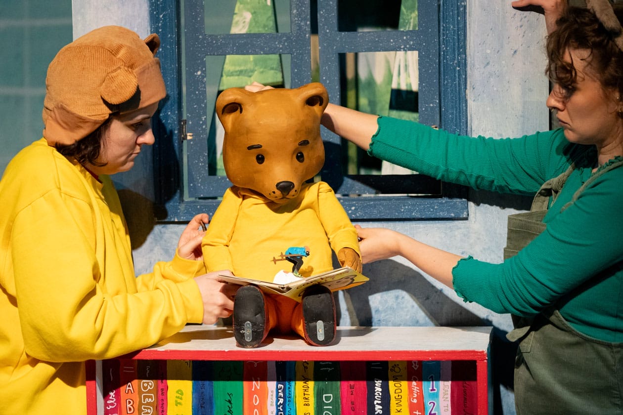 Two puppeteers hold a puppet of a bear in a yellow jumper reading a book. One of the puppeteers is dressed identically to the bear.
