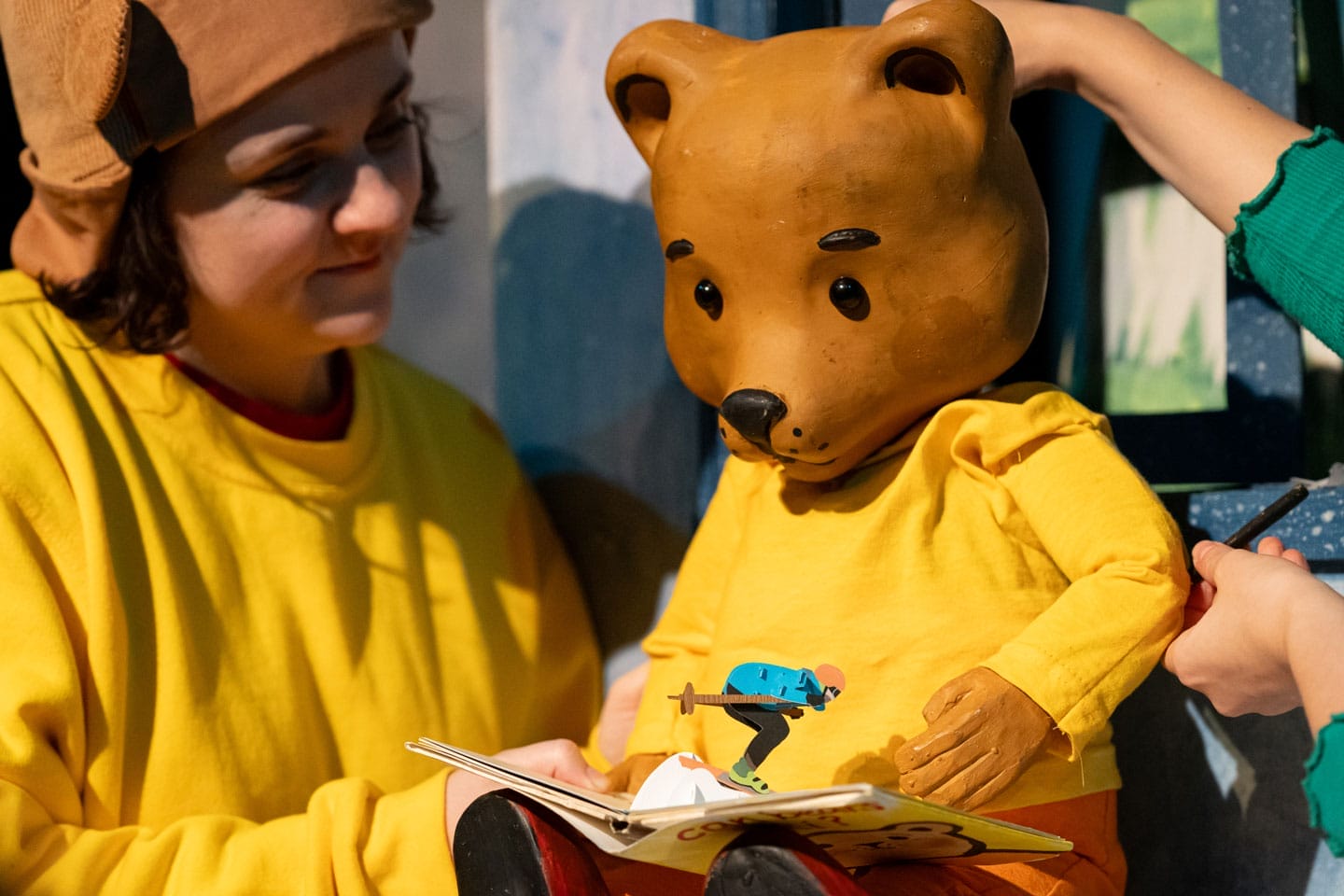 A close up of a bear puppet wearing a yellow jumper. Next to the bear, a puppeteer is wearing the same outfit.
