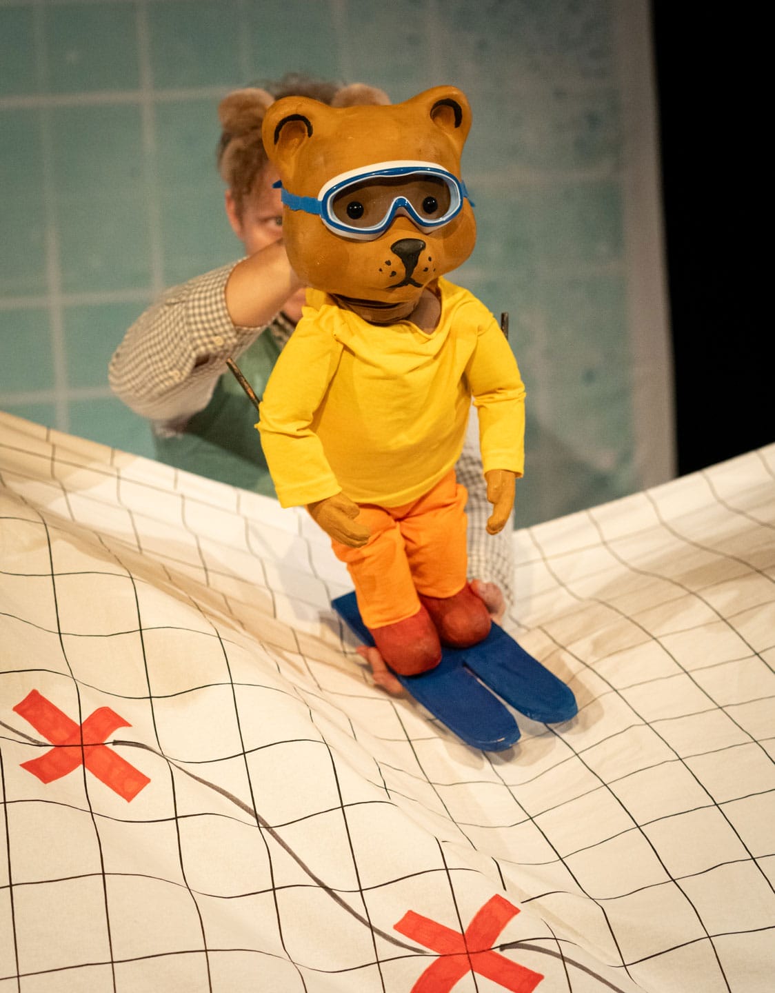 A puppet of a bear wearing a yellow jumper, orange trousers and goggles is sking on a paper landscape.
