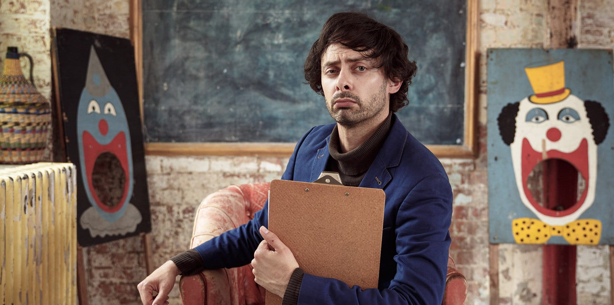 Man sits in a classroom pulling a funny face holding a clipboard