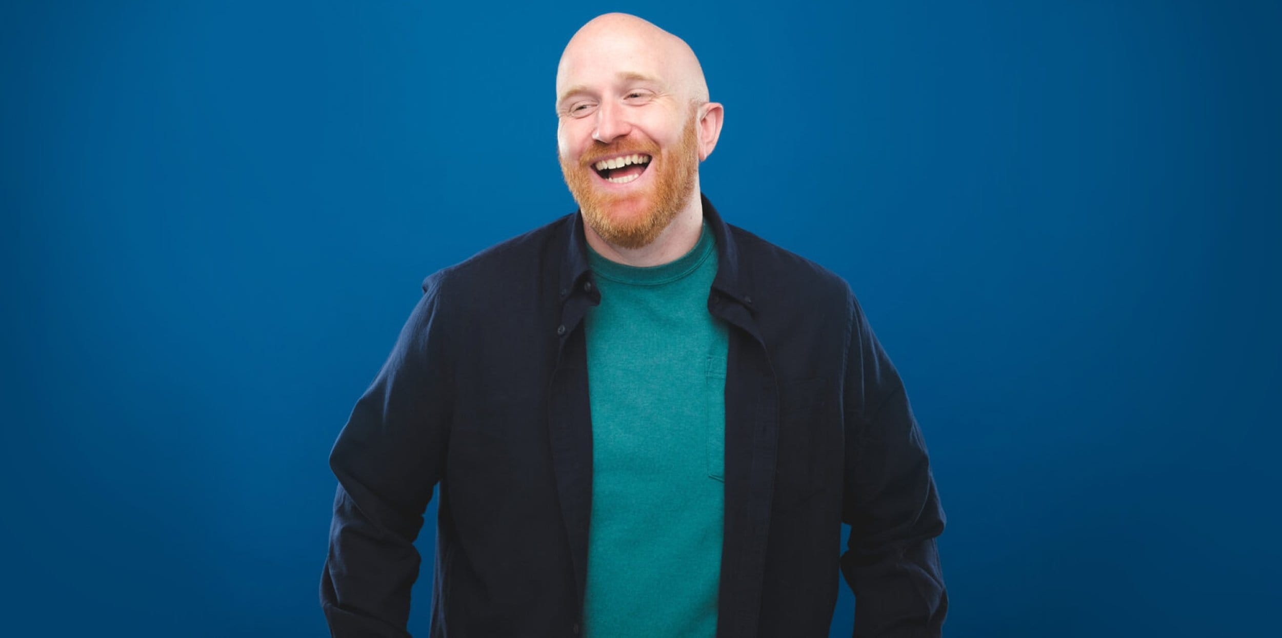 Comedian Ray Bradshaw Stand and laughs infront of a dark blue background