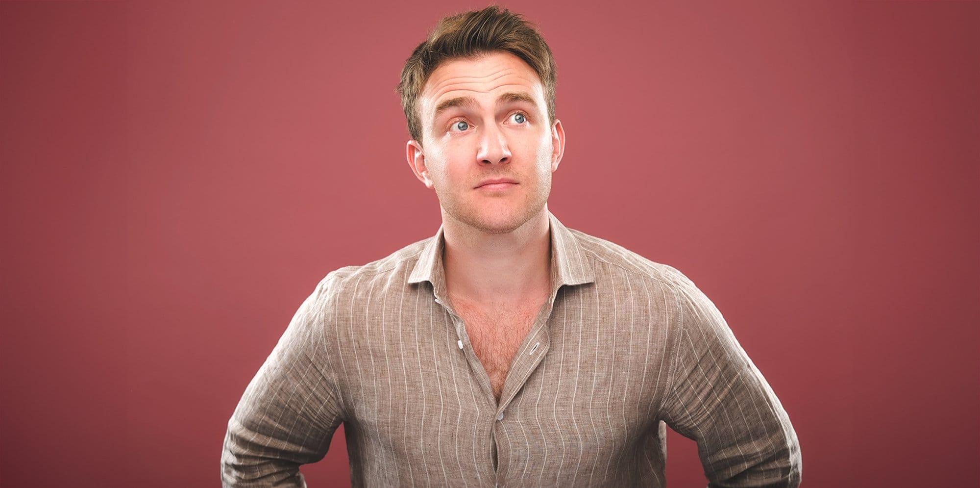 Tom Houghton, the comedian stands in a striped shirt on a light red background