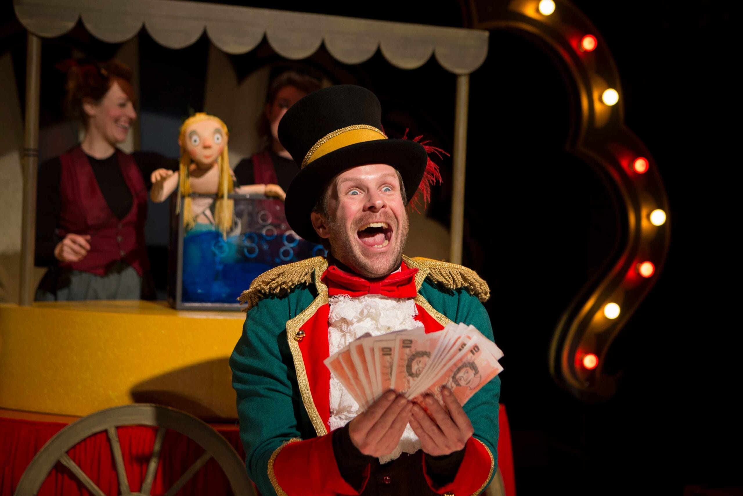 An actor dressed as Sam Sly holds up a lot of money looking elated. Behind him, the Singing Mermaid puppet sits in her tank. Two puppeteers can be seen in the background.