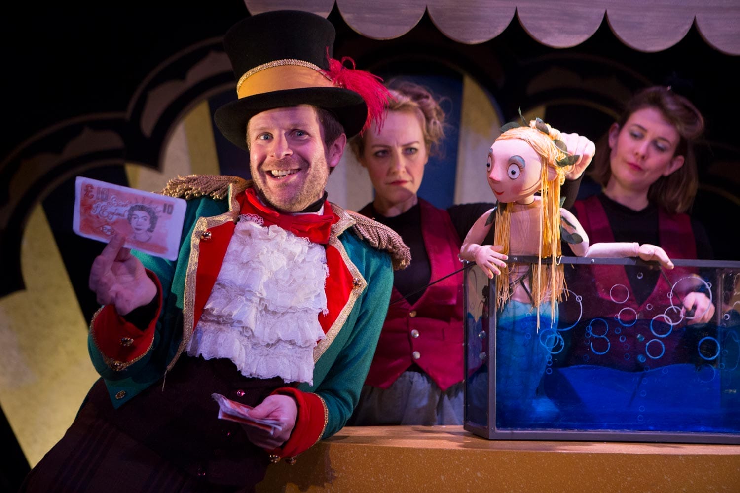 An actor dressed as Sam Sly holds up some money next to the Singing Mermaid puppet in her tank. Two puppeteers can be seen in the background.