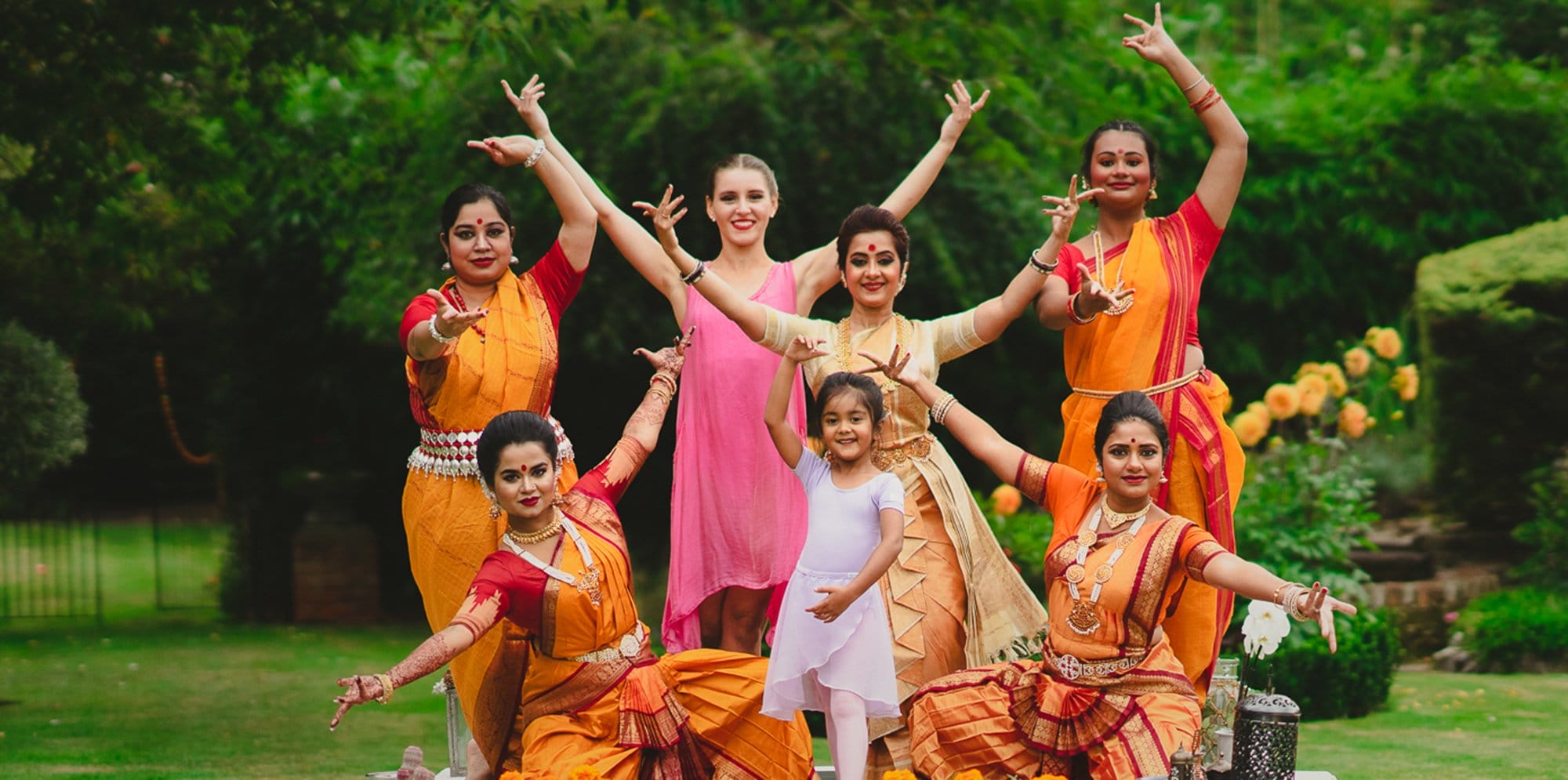 A group of women pose outdoors in traditional Bharatanatyam dance attire. The colour palette is vibrant red and orange. In the centre of the woman, a young child dancer smiles and poses.