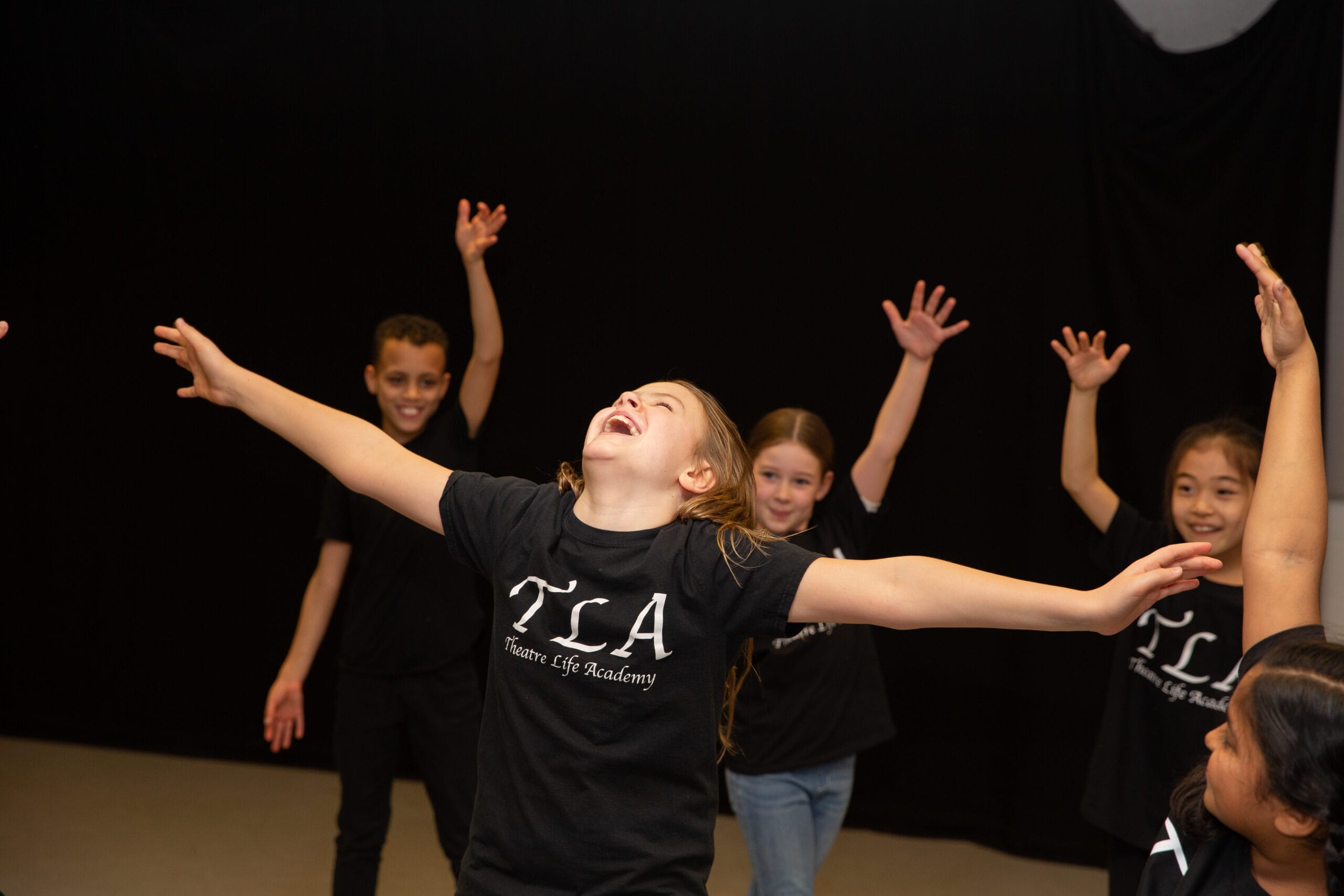 A young girl has her arms spread wide with a joyful expression as she engages in a dance class. Behind her, three other children are dancing happily with hands in the air.