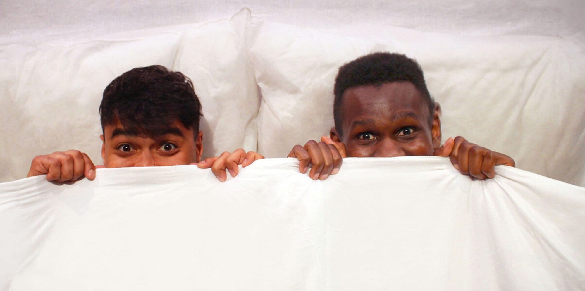 Two performers peek out from underneath the pillows and sheets with excited expressions.