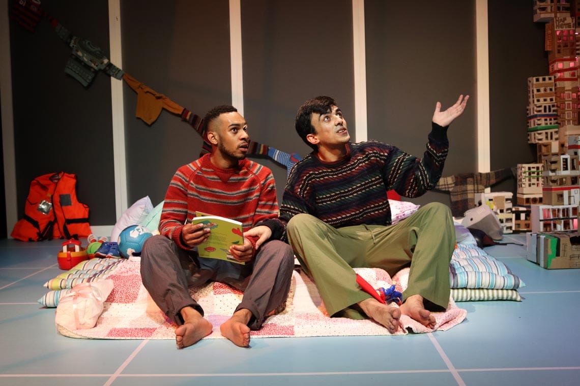 The two male characters sitting on their duvets and pillows looking up, talking looking inspired.