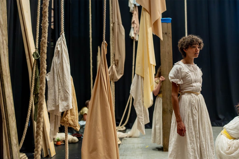 Rehearsal photography. We see the production set: dramatically draped sheets, cloths and ropes in beige and neutral tones. To the right, a performer stands, with a pensive expression.