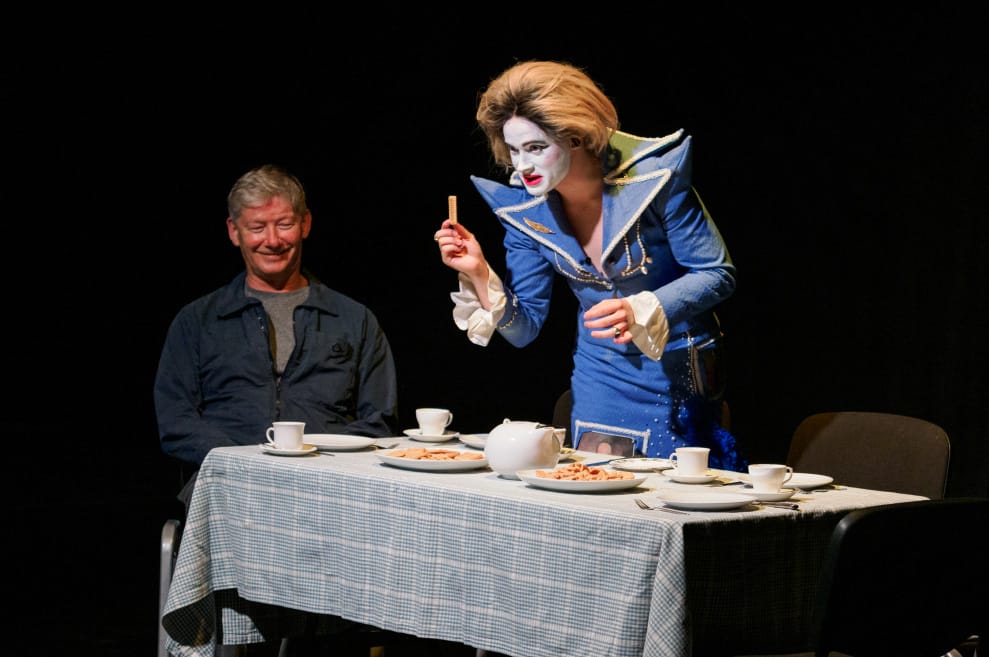 Performer wears dramatic drag costume as Margaret Thatcher. She commands a tea-party table, holding up a finger biscuit.