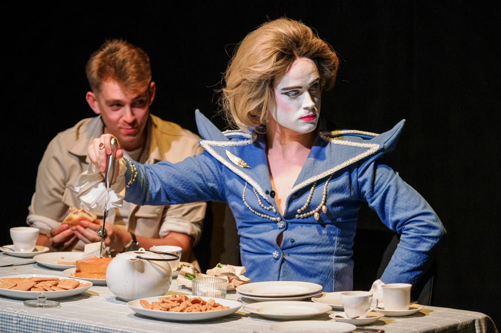 Performer dressed as Margaret Thatcher in dramatic wig, makeup and spiky blue blazer. She hosts a tea-party table, pronging a large knife into a victoria sponge cake.