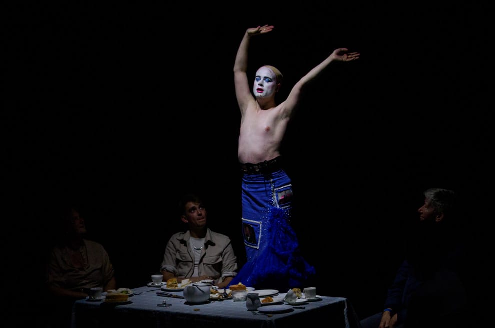 Performer stands at the tea-party table, arms waving in the air. Performer has removed wig and shirt, stripping down the layers of the Thatcher costume.
