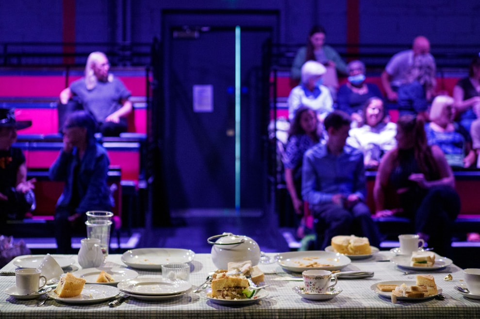 Photo is taken from the stage, showing the audience, blurred. In focus, we see the traditional British tea-party table. Teapots, sponge cake, cucumber, biscuits.