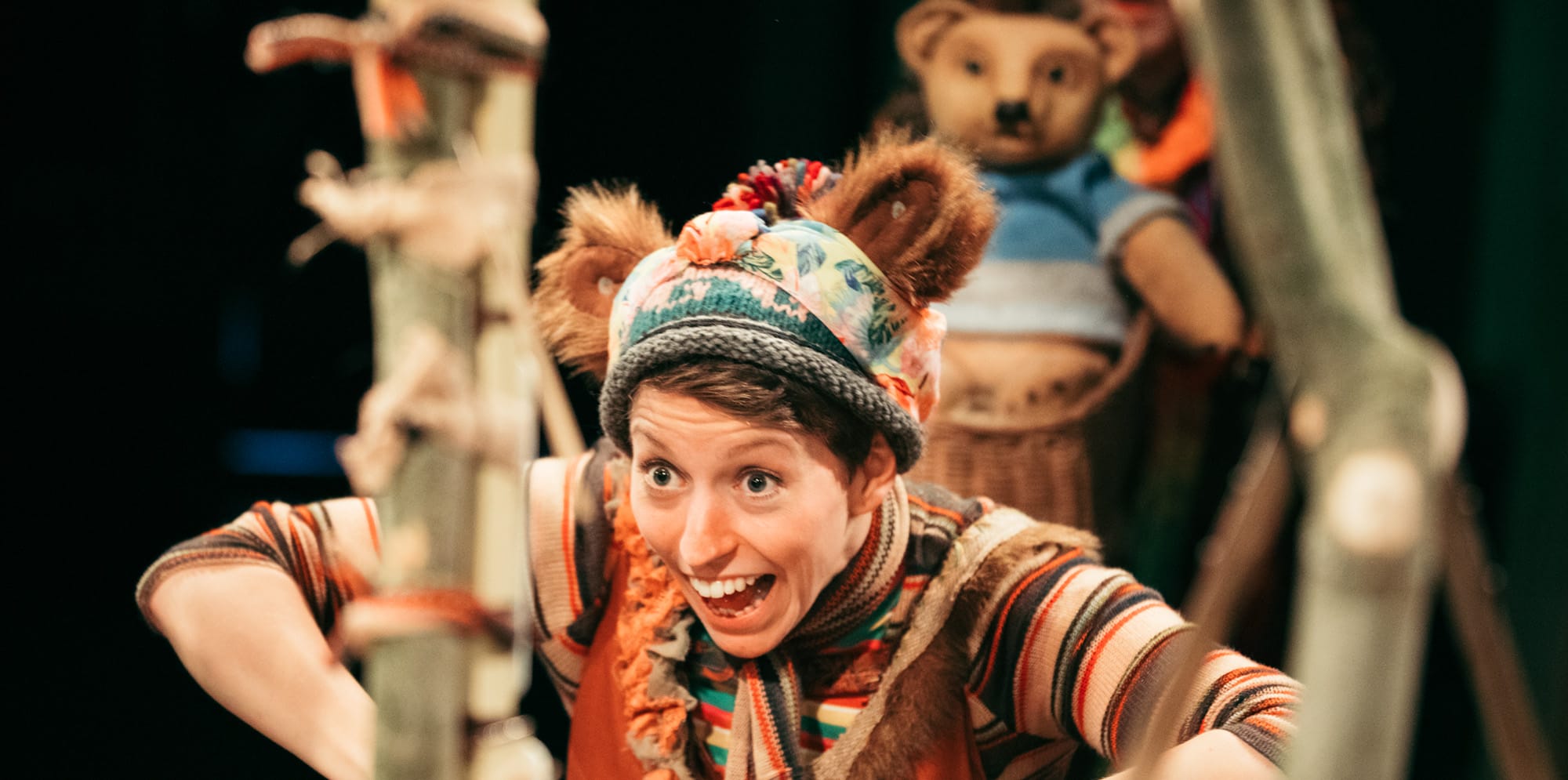 Performer wears fuzzy bear ears attached to a woolly hat, with a stripy shirt, scarf and furry collar. She has a big, wide-mouthed smile. Behind her, out of focus, a bear teddy is visible.