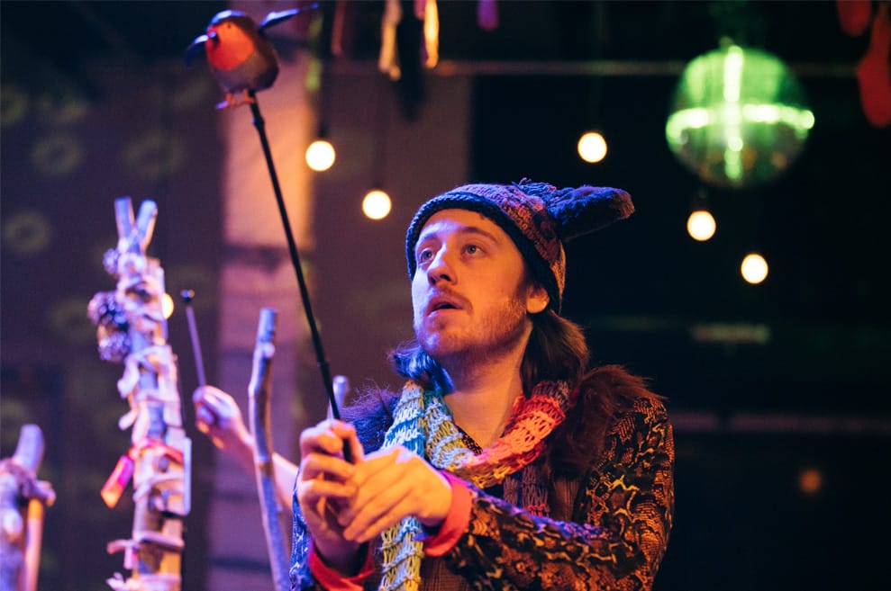 Performer wears assorted whimsical knitwear. He is holding a wooden robin puppet.