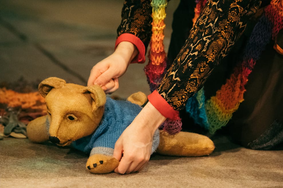 A little bear puppet crawls on the floor. The puppet is wearing a baby blue knitted jumper.