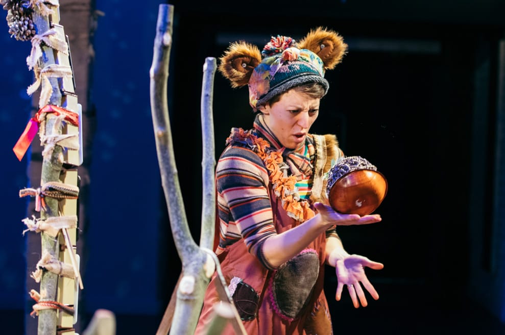 Performer wears knitted hat with fuzzy bear ears, colourful patterns and rust-orange dungarees. She is holding a very large wooden acorn.