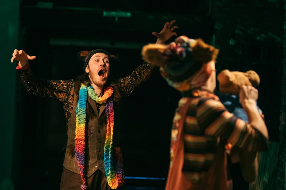 Two performers interact onstage. One has their back to camera, the other is visible with arms outstretched and mouth wide open. He is wearing a rainbow stripy knitted scarf.