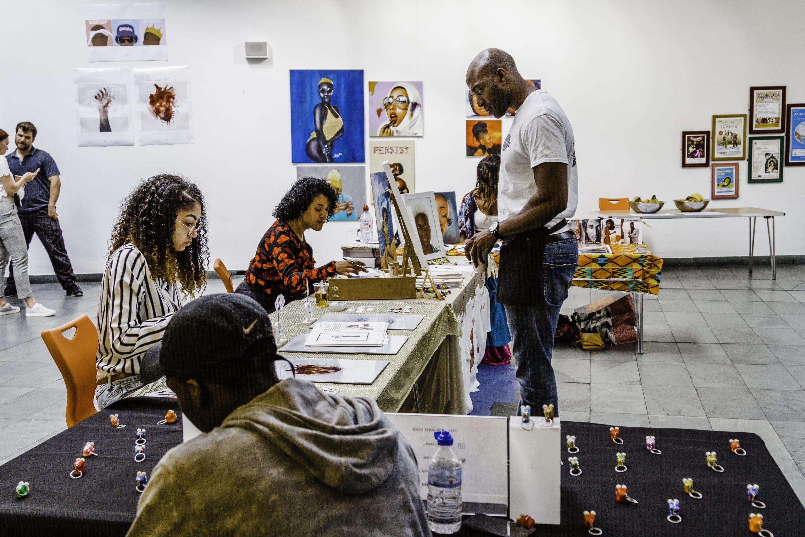 The artsdepot building is busy during an art event. In the centre of the image, black-owned arts and crafts vendors display their work. Vibrant paintings, prints, t-shirts and keyrings are on display as customers engage with the stalls.