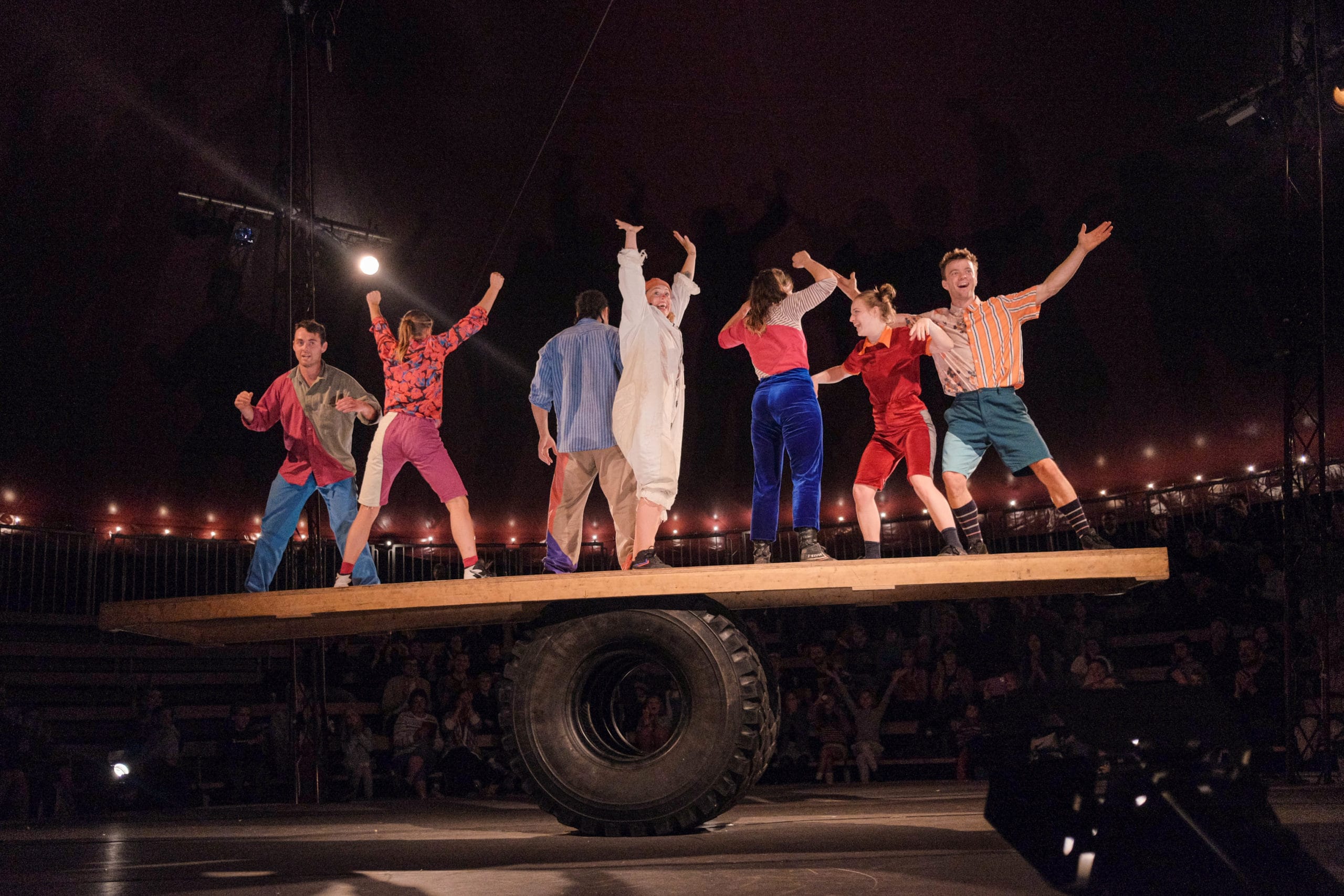 7 Revel Puck circus performers balance on top of a wooden seesaw, held up by an extra large large rubber tyre. They carefully balance to suspend the seesaw at an exact equilibrium. They are all wearing vibrant assorted circus-style costumes.