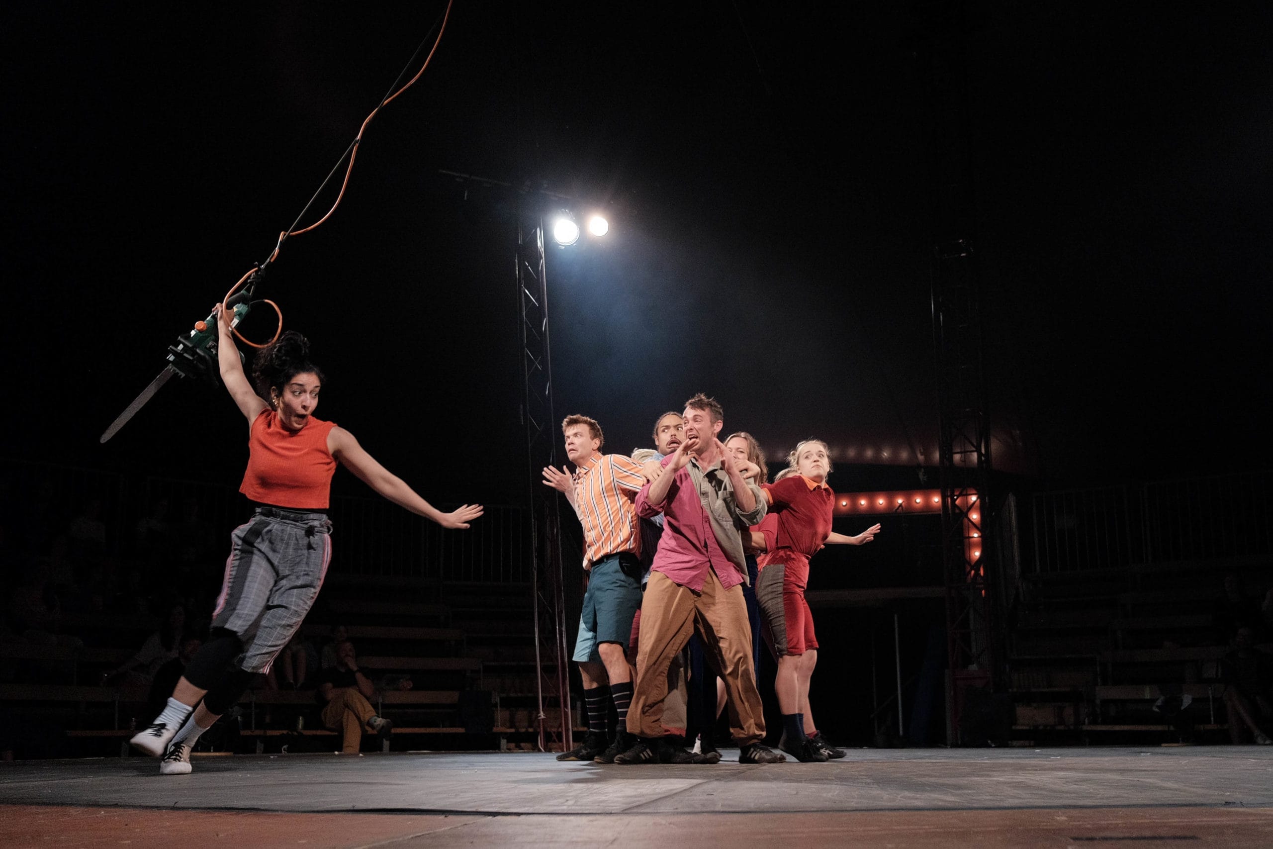 Several Revel Puck circus members are grouped in the centre of the stage, wearing dramatic expressions. One member of the troupe is attached to a suspended rope and chainsaw, about to take flight.