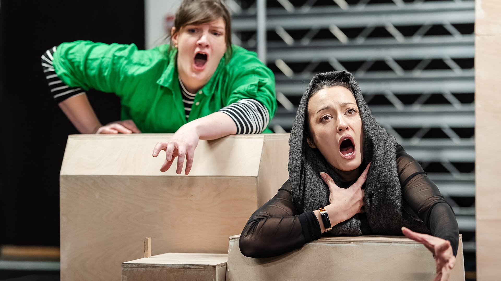 Two performers lean over low level cardboard boxes whilst singing.