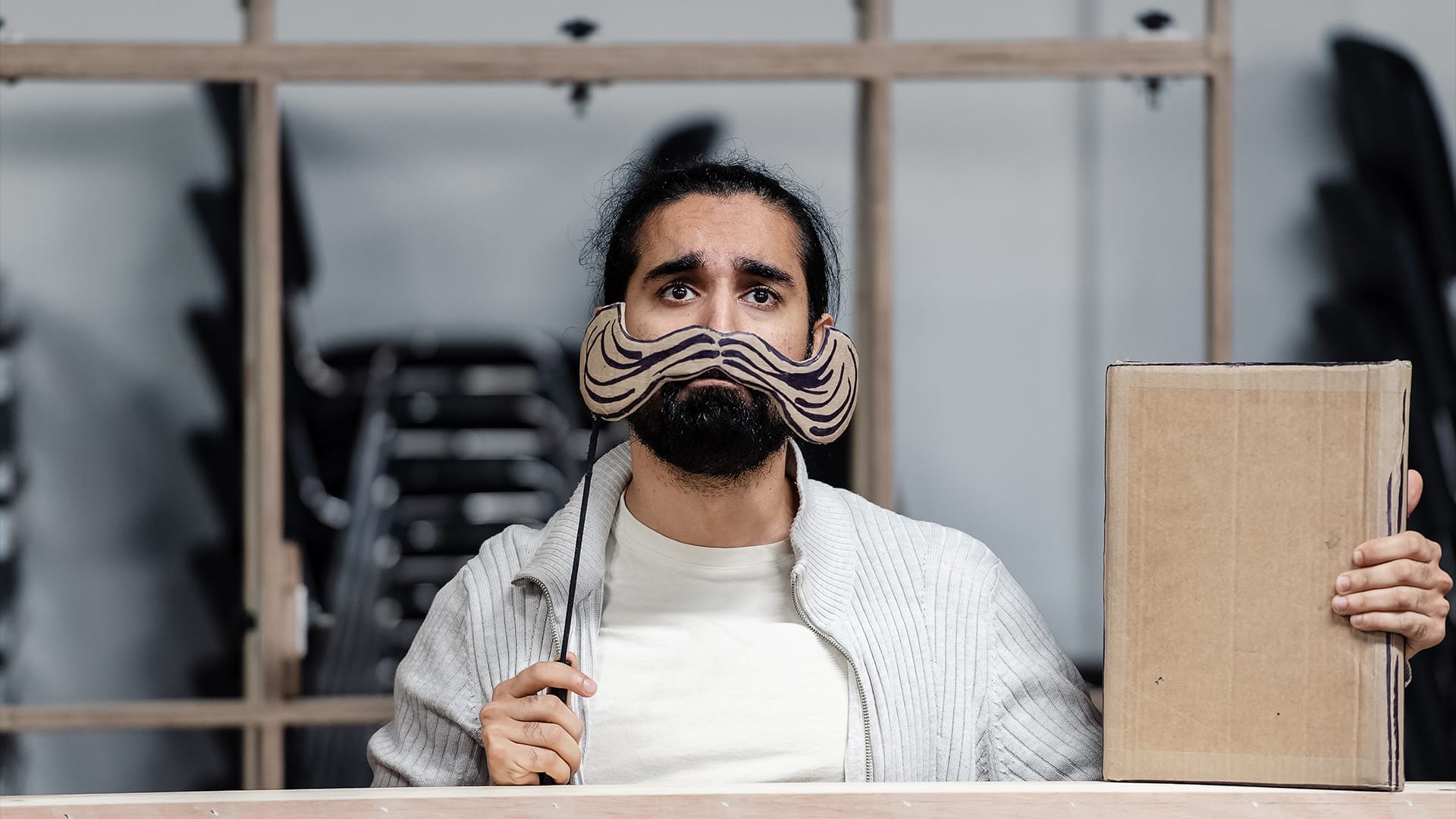 A performer holds a 2D cardboard book in one hand, and holds a big twirly Victorian style cardboard moustache in front of his face with the other.
