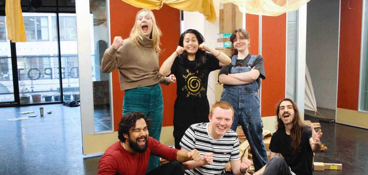 Six performers from artsdepot's 2023/2024 artist residencies pose in the artsdepot creation space. Three performers stand whilst three crouch in front. They are all smiling with their hands in dynamic, cheerful poses.