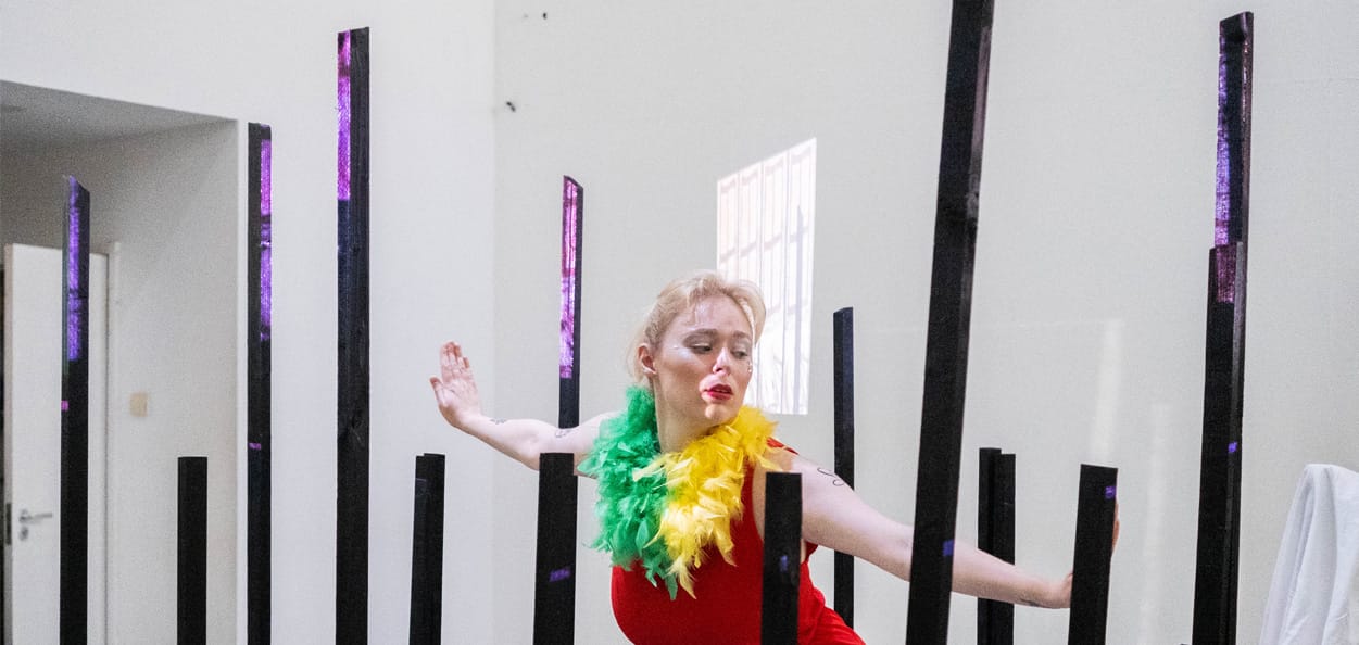 A performance art piece in a white studio space. Dark metal bars frame a performer, posing in the centre. The performer is a white woman with blonde hair; she is wearing a red jumpsuit, red lipstick and a green and yellow neck piece.