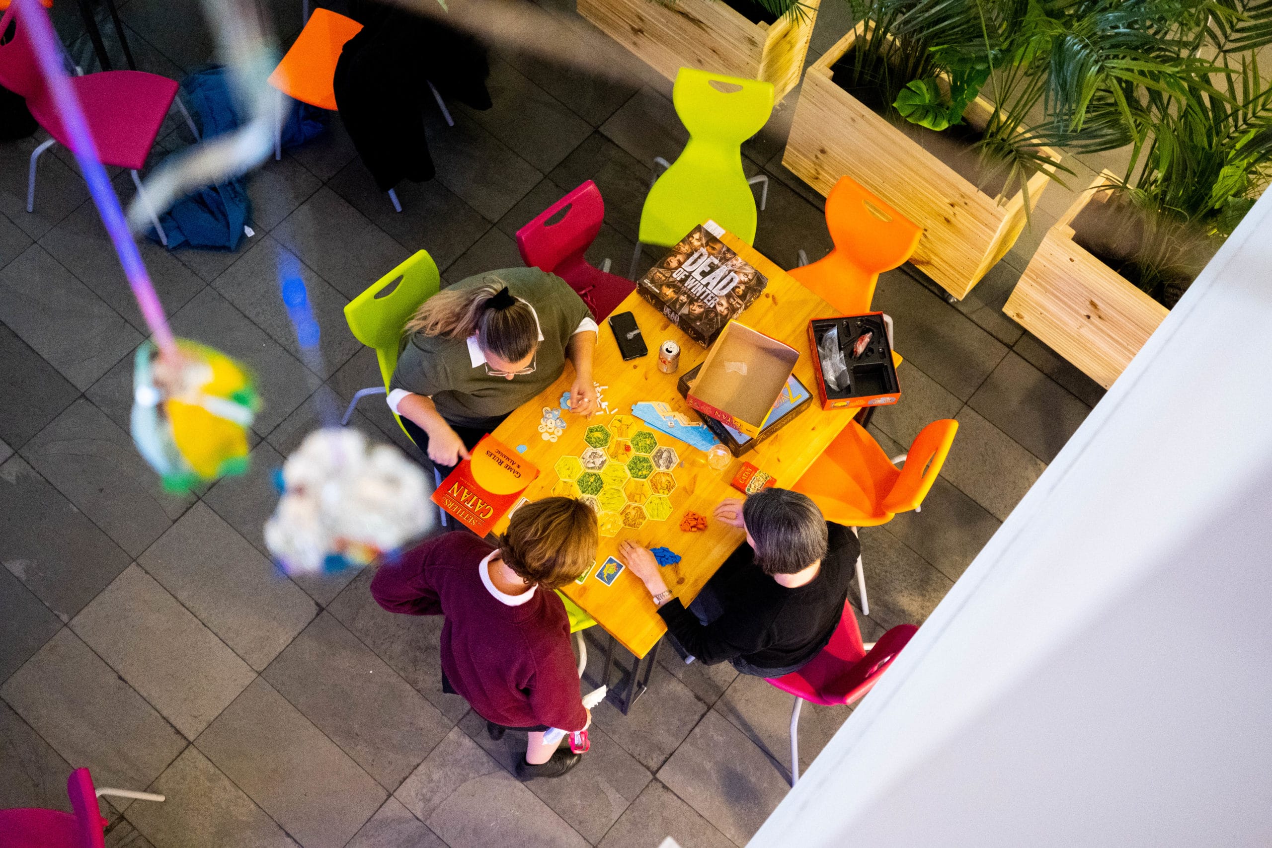 Birds eye view of the artsdepot cafe area during an evening game night. Three young people are playing a colourful board game whilst chatting.