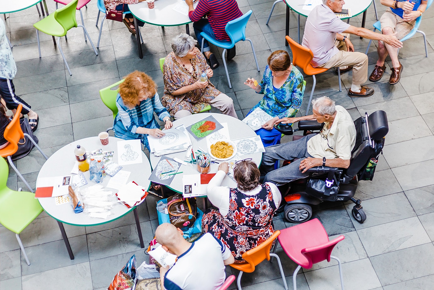Bird's eye view of the artsdepot cafe. A group of older people are participating in an art activity. They sit on colourful chairs with a large bowl of nachos in the centre of the table.